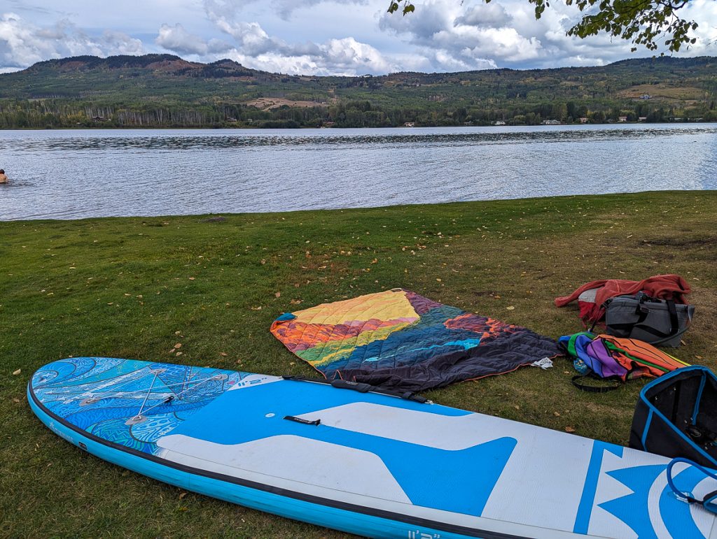 A SUP lying on the grass next to a blanket and other bags, with a body of water and more land in the background.