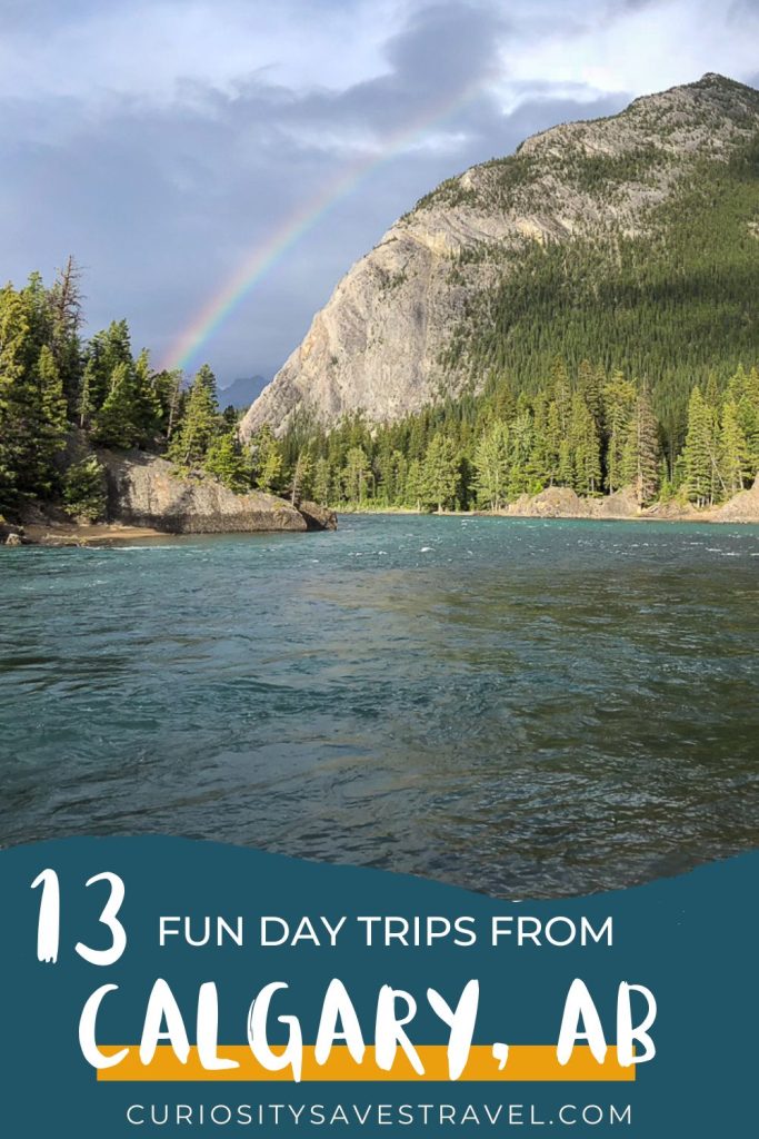 A river next to a wooded mountain with a rainbow in the background and a text box at the bottom which reads "13 fun day trips from Calgary, AB."
