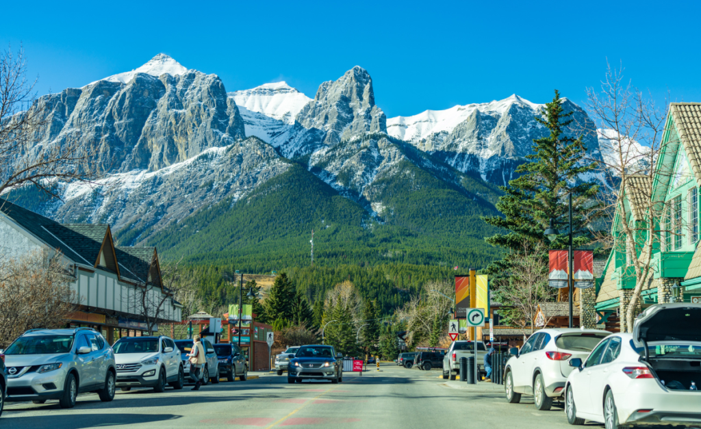 A road lined with parked cars and shops leading towards a mountain with green trees at the bottom and snow at the peaks with blue sky behind.