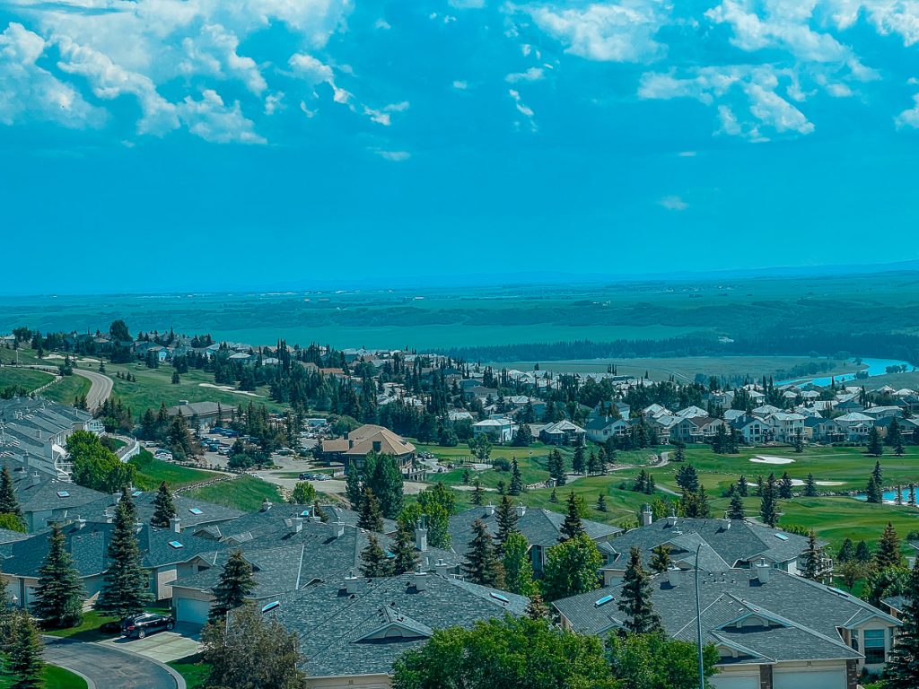 A view across lots of houses and buildings on a green landscape with trees dotted throughout towards a wide open landscape and blue sky above.