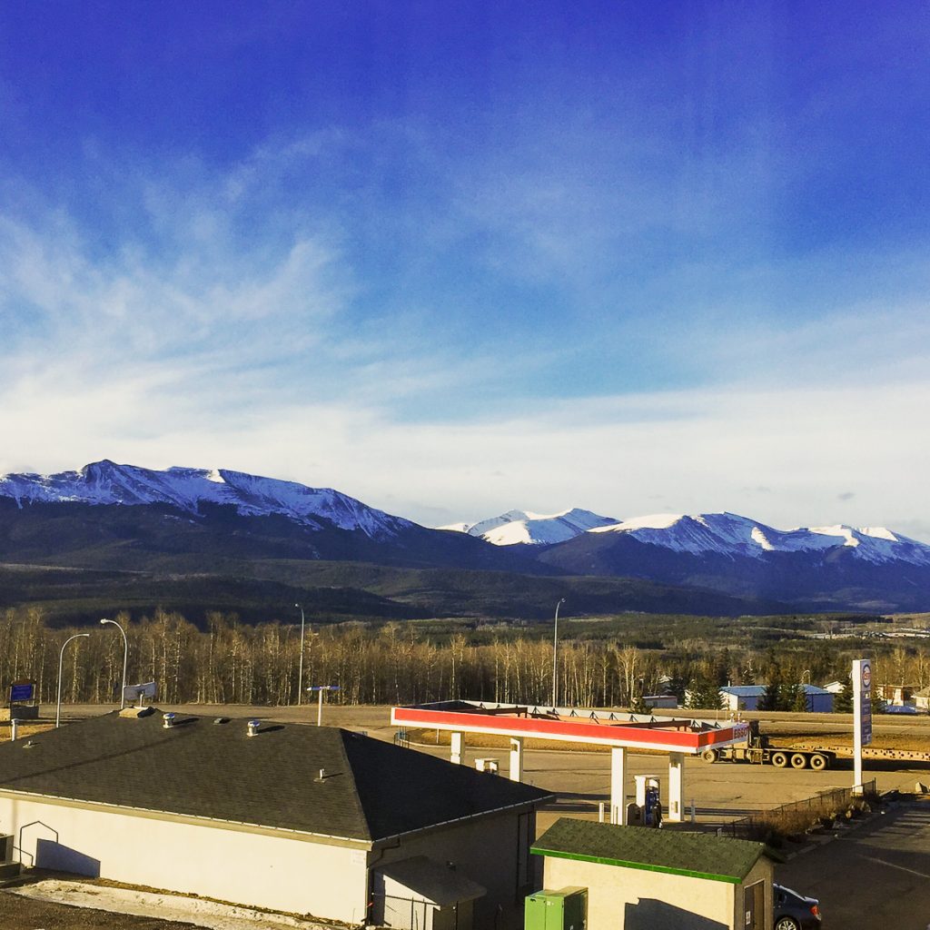 A view across a building rooftop and gas station towards forest and snow-topped mountains.