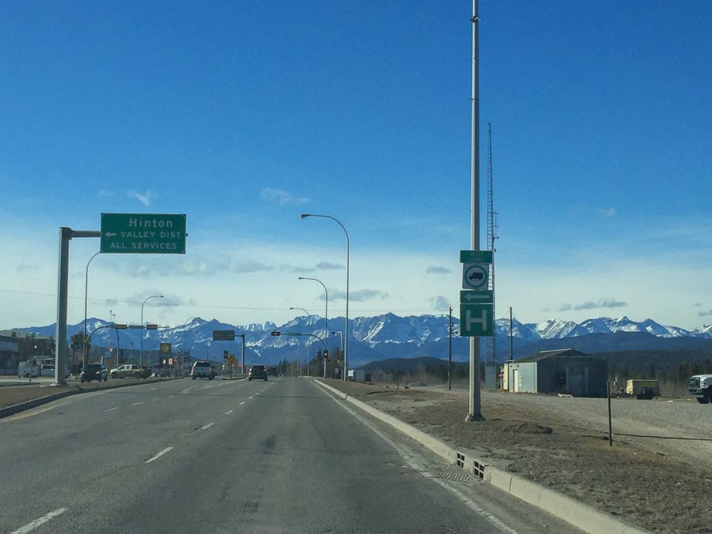 A highway with cars on it and a sign pointing to Hinton with snow-capped mountains and blue sky in the distance.