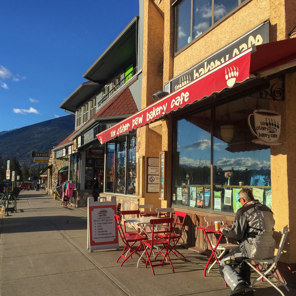 A shop on a shop-lined street with a red sign that reads "The Other Paw Bakery Cafe" with a man sitting with his back to the camera at an outdoor table with a coffee.
