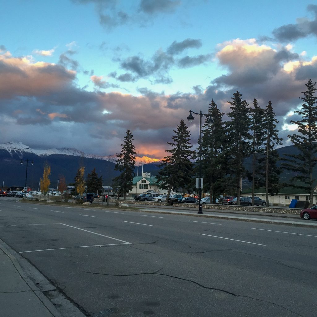 A view across a road to a carpark with a fiery sunset just peeking over a snowy mountaintop in the distance with fluffy clouds above.