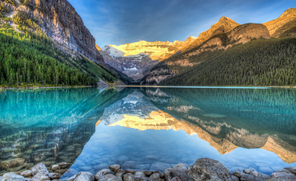 A photo of a very still lake that's perfectly reflecting the mountains and trees on the shores with blue sky above.