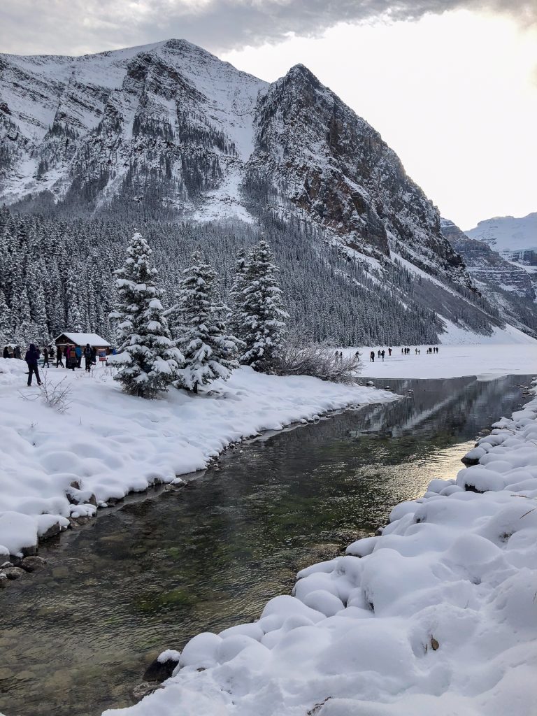 A snowy river with people and a small wooden building on the banks and a mountain in the background.