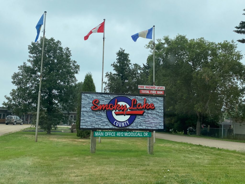 A roadside sign that says "Smoky Lake County" on some grass with three flag poles and trees behind it.