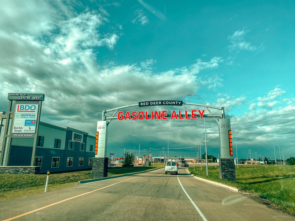 A sign across a road that says "Red Deer County Gasoline Alley" with some cars and buildings in the distance.