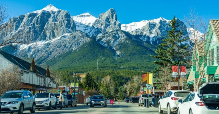 A road lined with parked cars and shops leading towards a mountain with green trees at the bottom and snow at the peaks with blue sky behind.