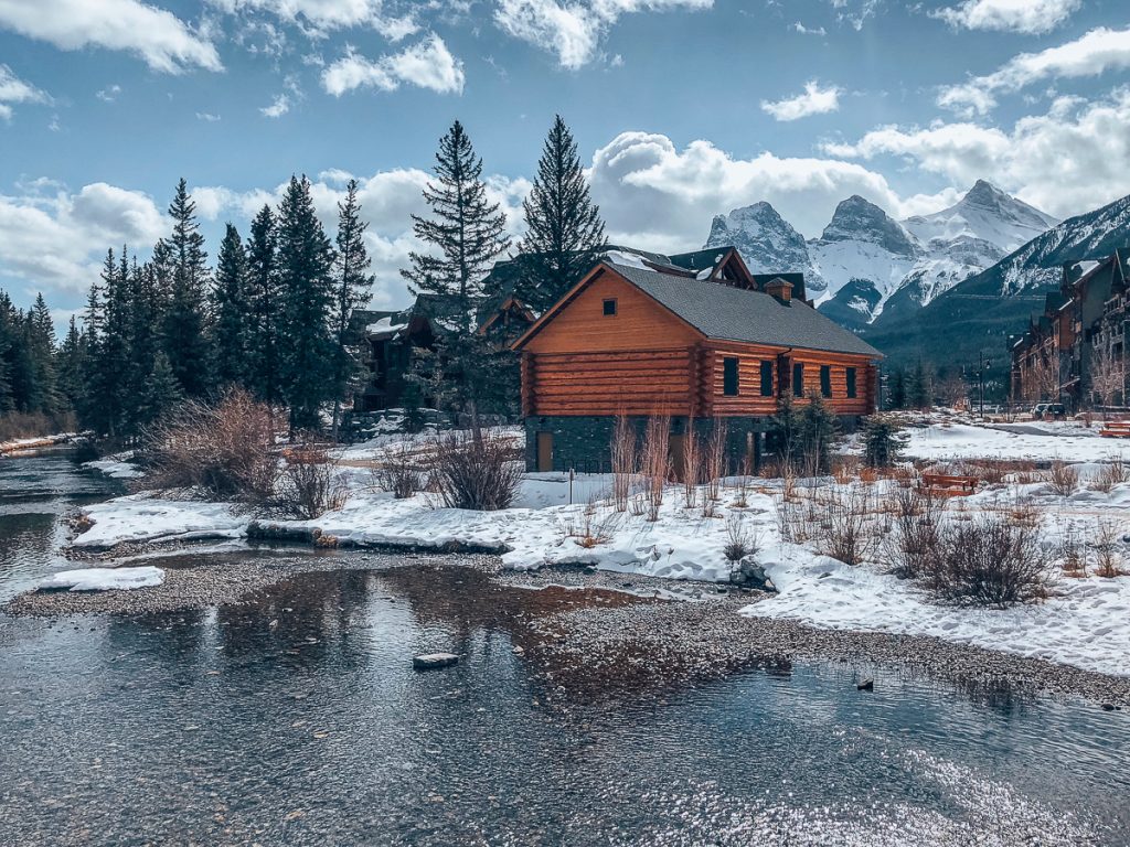 A wooden log cabin next to a river with snow on the ground, surrounded by pine trees and with snowy mountains in the background.