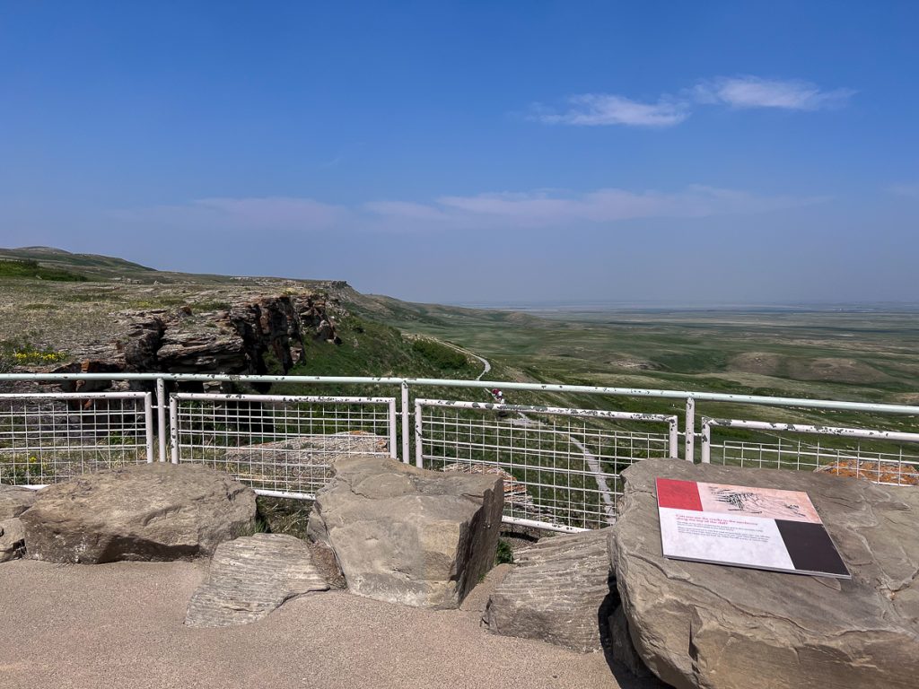 A lookout with rocks, a fence and an information board looking out over cliffs and a green plain with a blue sky overhead.