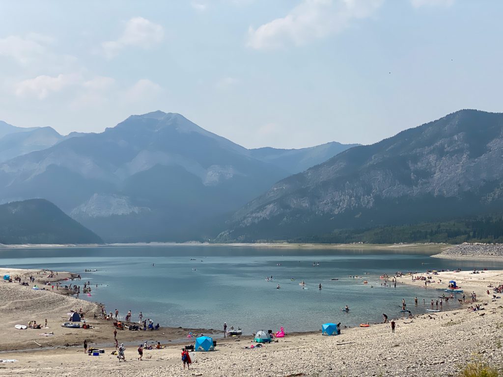 A rocky beach next to a lake with lots of people at the edge and in the water, with large mountains in the background.