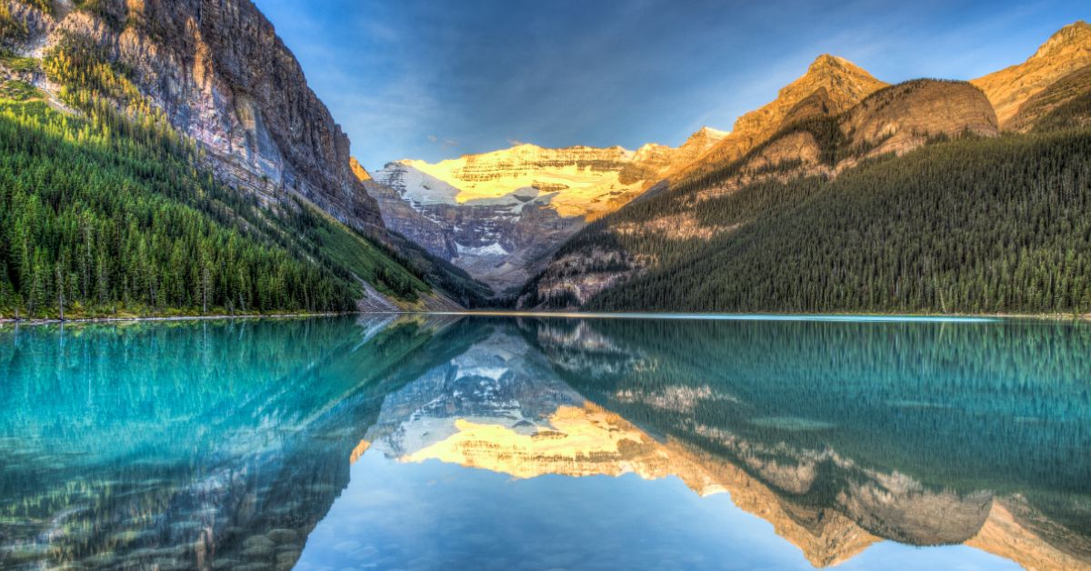A photo of a very still lake that's perfectly reflecting the mountains and trees on the shores with blue sky above.
