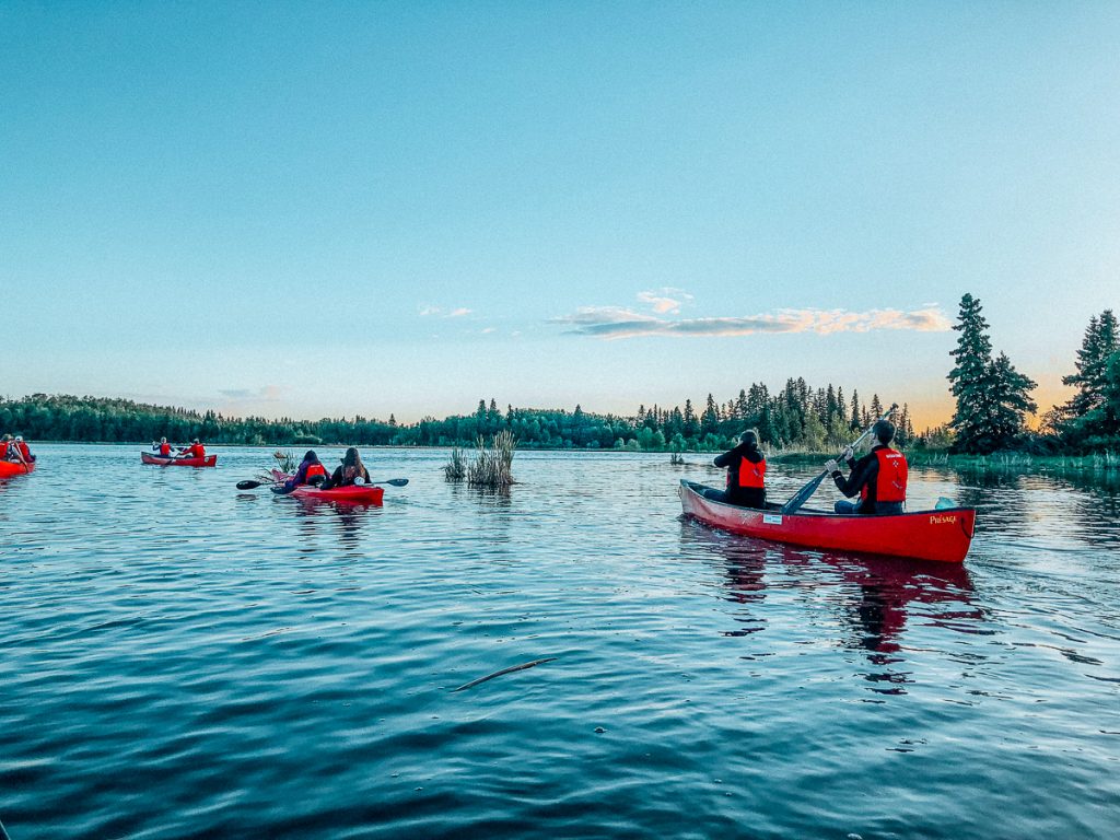 A landscape photo of six people kayaking on a lake in three red kayaks with clear blue skies above and some trees on the landline in the distance.