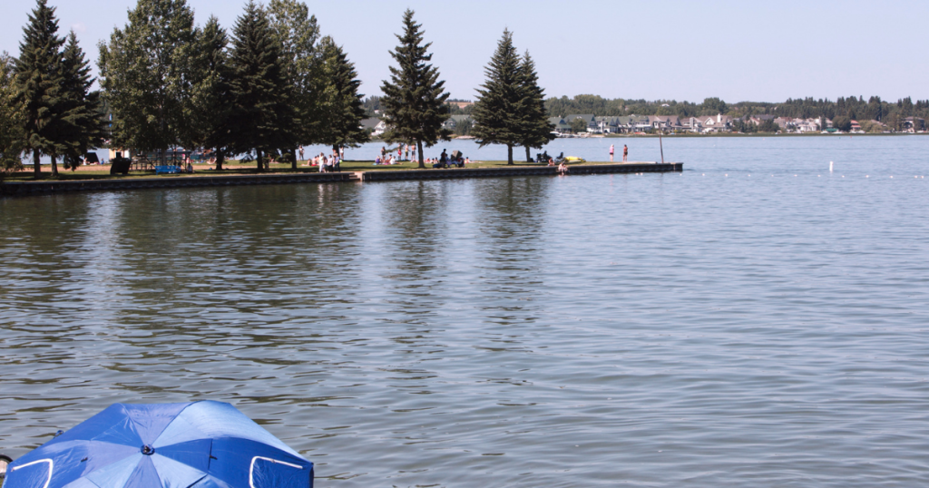 A view across a lake to a grassy area with trees and people, with more trees and houses in the far distance.