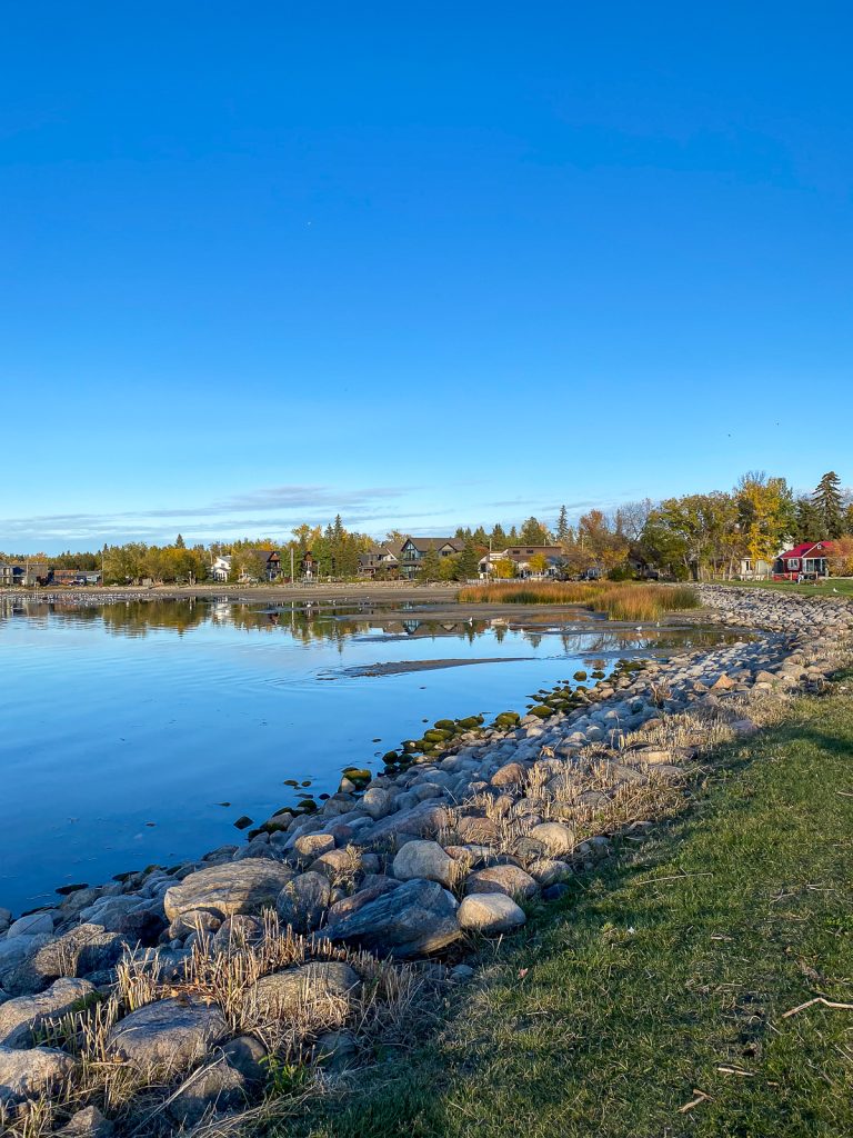A rocky lake curving away from the photographer to houses and trees with a clear blue sky above.