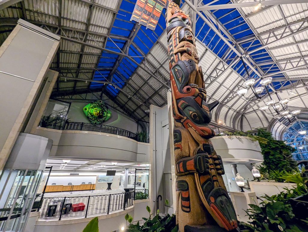 A huge wooden totem inside a convention center-type building with high ceilings, balconies and plant life around.