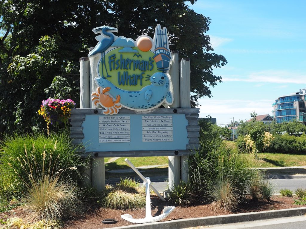 A wooden sign that reads Fisherman's Wharf with 3D images of a crane, a boat, a crab and a seal. Underneath are directions to places of interest and an anchor lying on the ground.