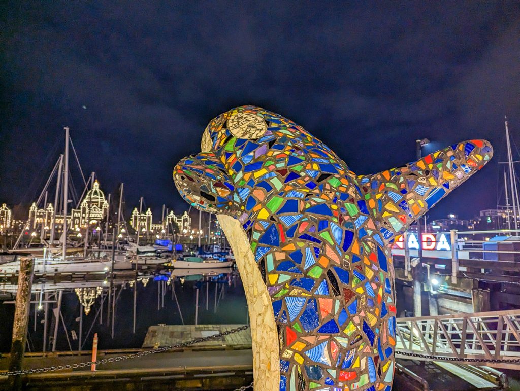 A statue of an orca made from colored glass leaping in front of a marina at night.
