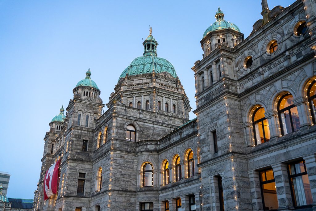 A grand stone building with lights dotted all over the exterior, green domed roofs and a large Canadian flag hanging on the exterior.