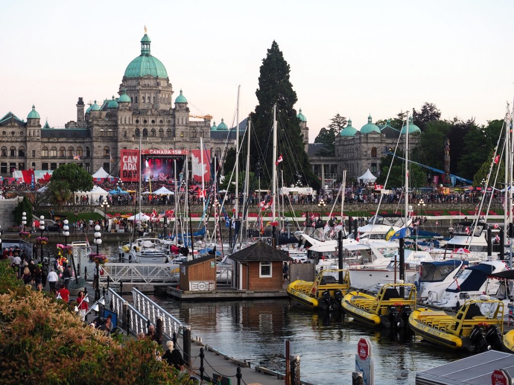 A harbor with boats docked next to a grand stone building where a stage is set up and people are watching a performance with Canadian flags all over the place.