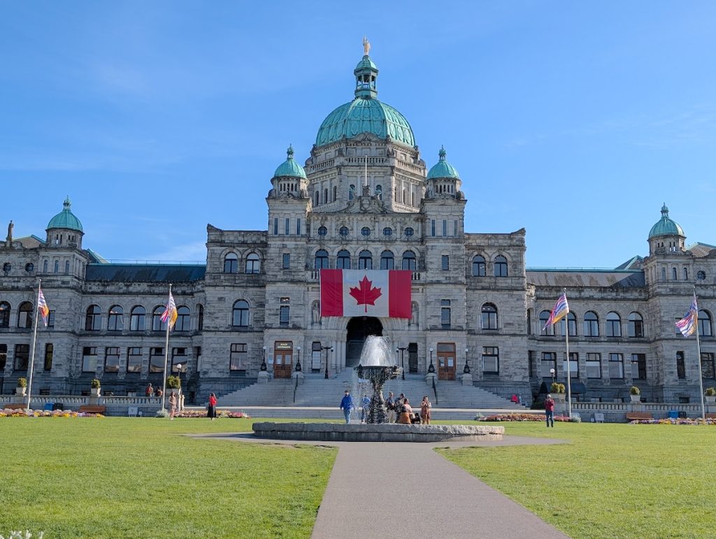 A palatial stone building with multiple domed roofs,  a water fountain out the front and a large Canadian flag hanging from the building on a sunny day.