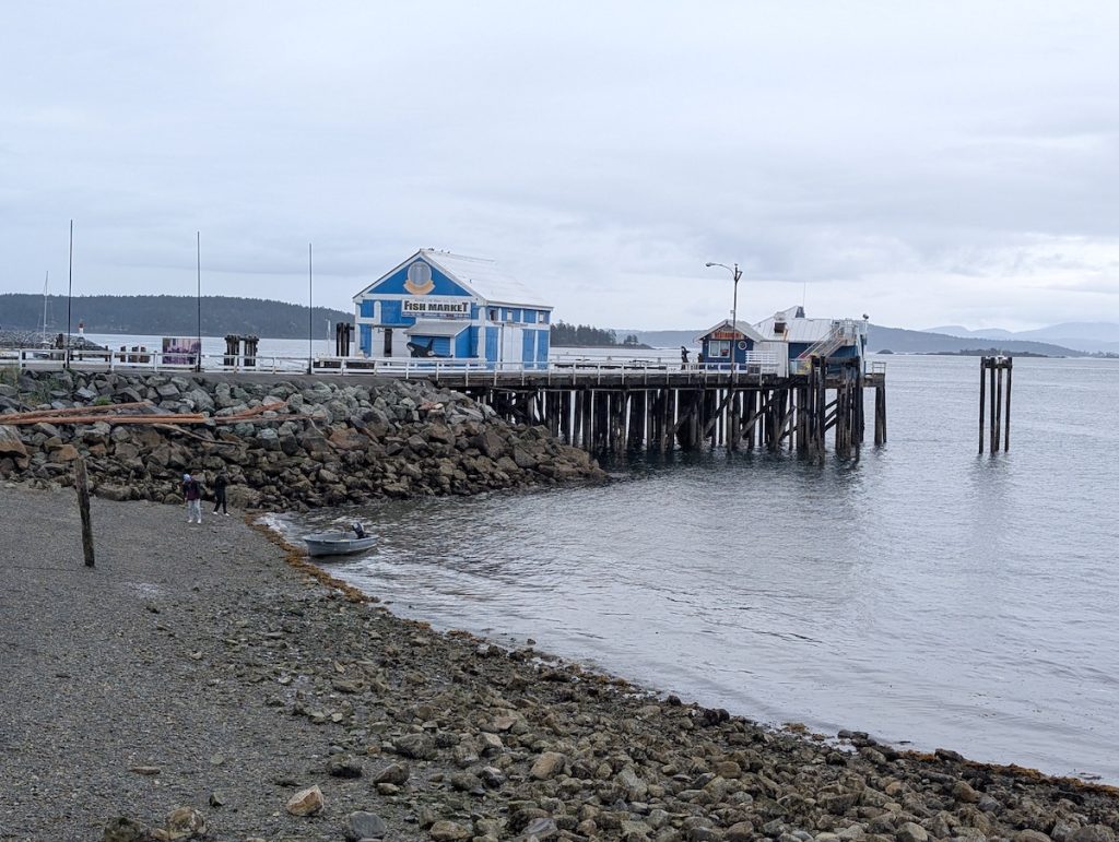 A rocky beach and jetty with a blue and white wooden building on it that says Fish Market on an overcast day.
