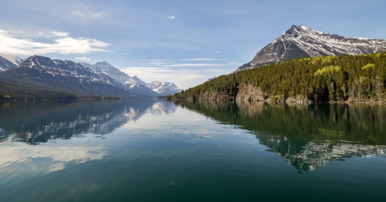 A calm lake reflecting trees and snow-capped mountains on both sides with blue skies and some white clouds above.