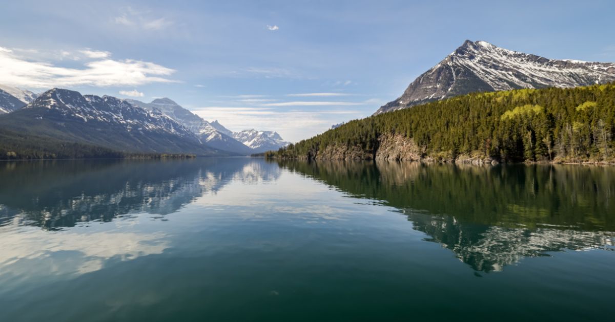 A calm lake reflecting trees and snow-capped mountains on both sides with blue skies and some white clouds above.