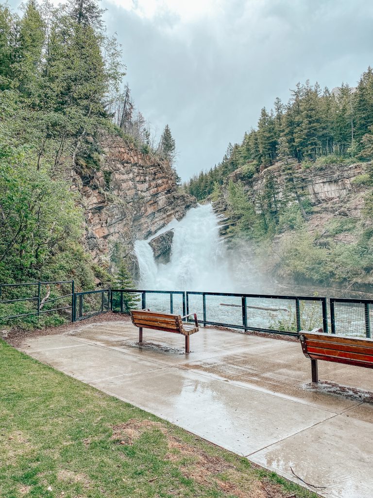 Some wooden benches on cement next to a railing overlooking a waterfall.
