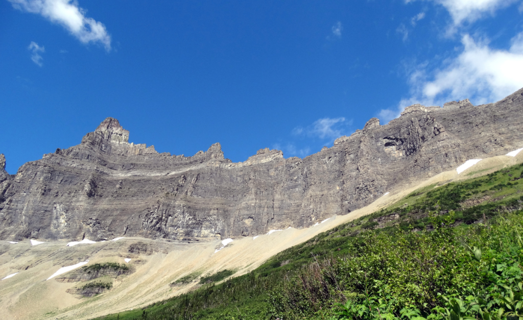 A rocky mountain with striations rising above a green slope with blue sky above.
