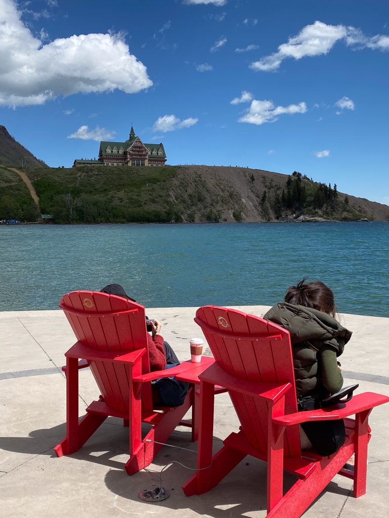 Two people seen from behind sitting in red wooden chairs next to a lake with a building on a headland across the water.