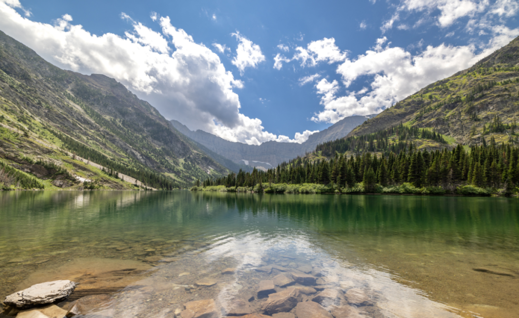 A lake with clear water showing rocks underneath with forested mountains in the distance and a blue sky with fluffy white clouds.