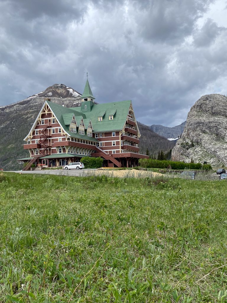 Green grass in front of a red and cream wooden building with a green roof, with mountains and a cloudy sky behind.