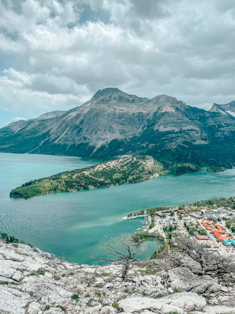 A view from high up on a rocky outcrop looking down on a small town next to a large lake with more land jutting out on the other side and another rocky mountain.