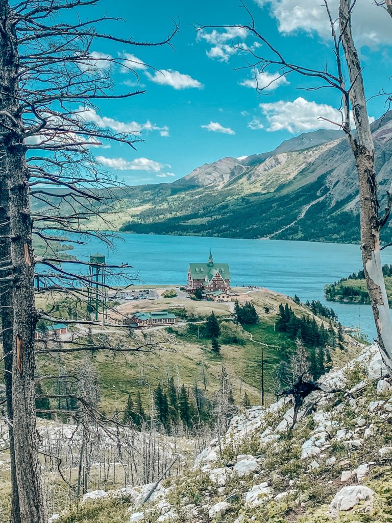 A view from a hill down to a hotel on an outcrop overlooking a bright blue lake with more mountains and blue sky in the background.