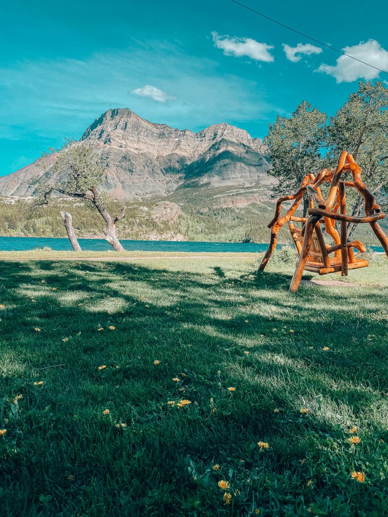 A wooden swing on a grassy area next to water with mountains in the background.