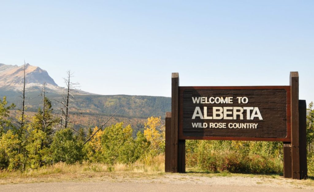A wooden sign on the side of the road that reads "Welcome to Alberta Wild Rose Country" with trees and hills in the background.