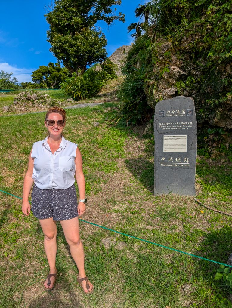 Sooz wearing shorts and a white sleeveless blouse smiling while standing next to a stone pillar with information in Japanese and English.