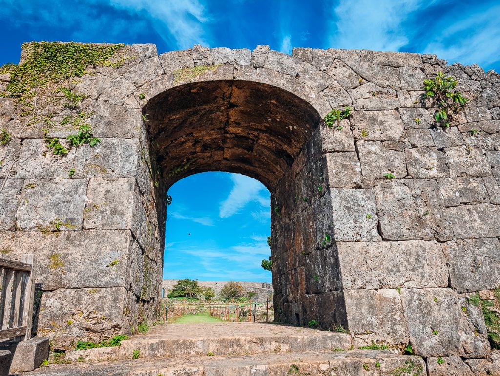 An archway in a stone wall leading towards more stone walls and a blue sky.