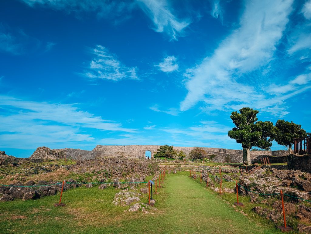 A fenced off series of rocks in a green field with a stone wall in the distance with an arched doorway and blue skies above.