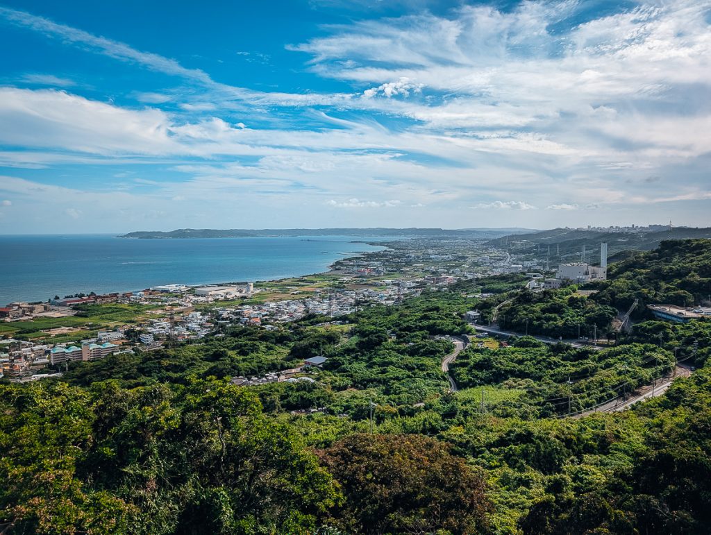 An aerial view over green trees and roads leading to a coastal city with blue skies and white clouds above.