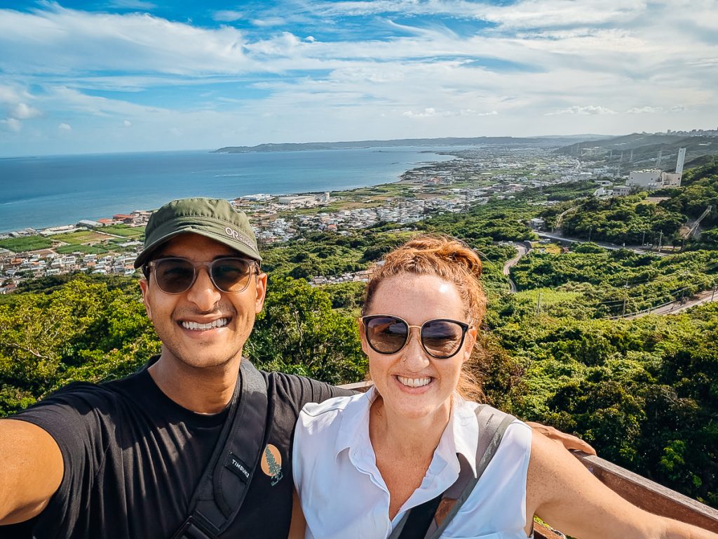 Ganesh and Sooz posing for a selfie at a viewpoint with greenery and roads behind them leading to a coastal city.