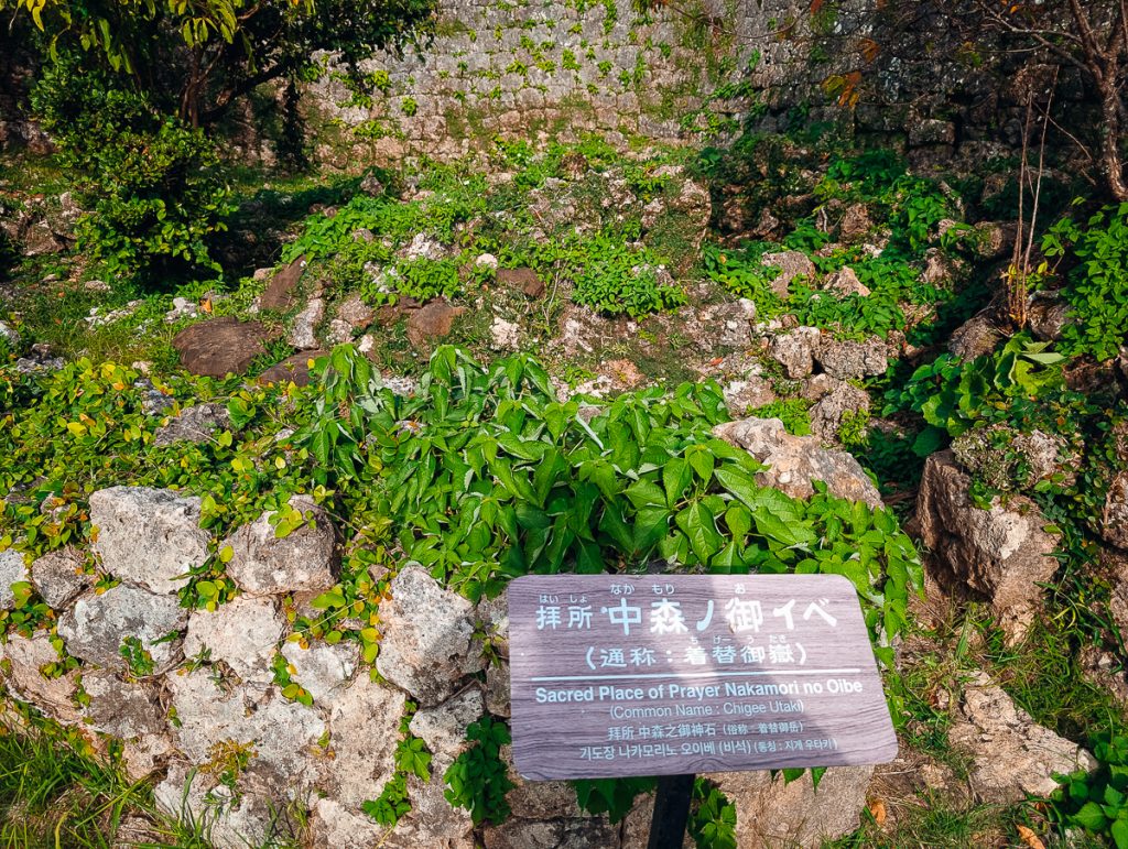 A wooden sign in front of rocks and plants with text in English and Japanese saying it's a sacred place of prayer Nakamori no Oibe.