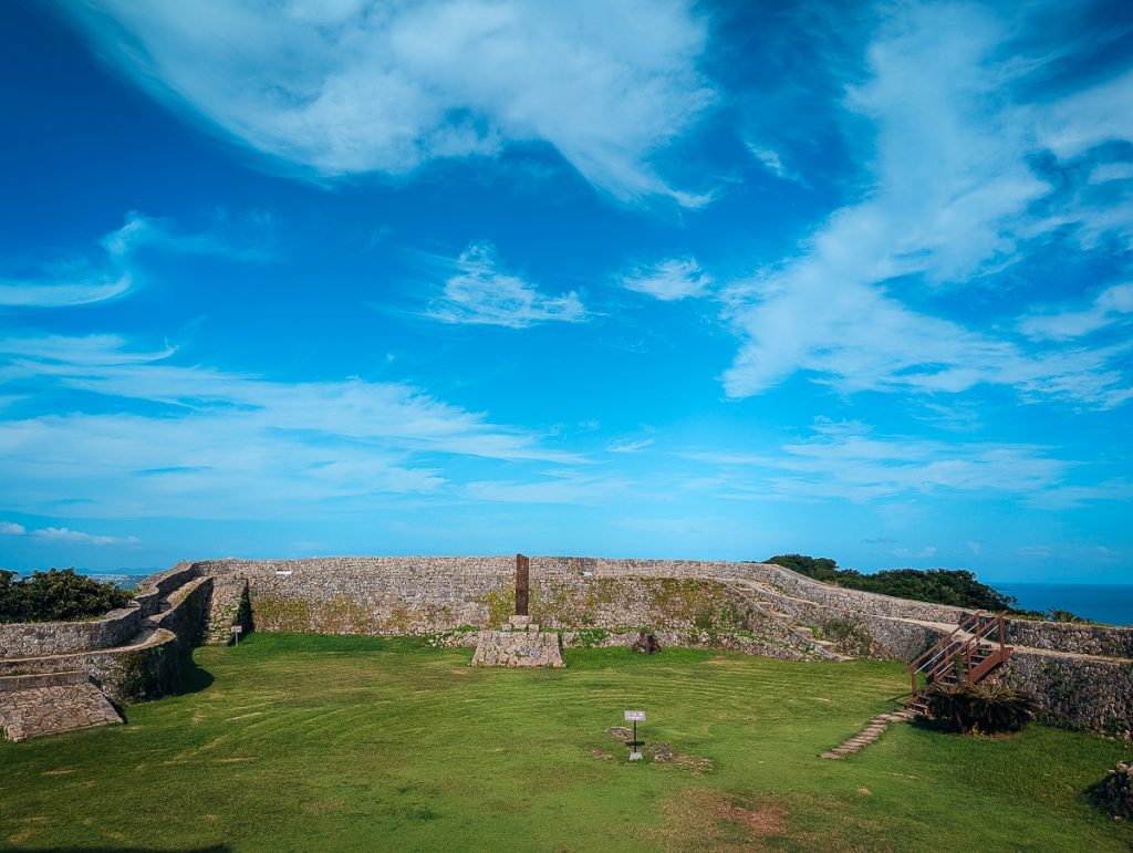 A curved stone wall next to green grass and a pillar on a stone structure with blue skies above.