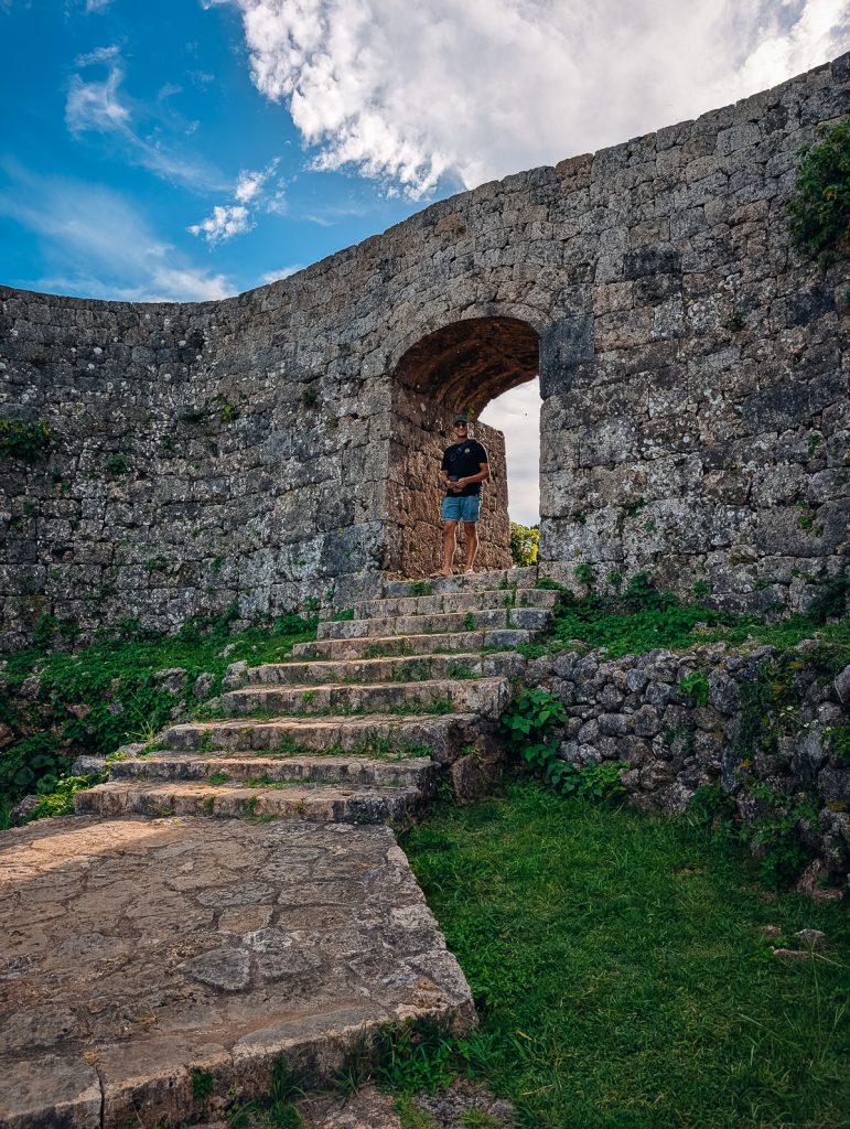 A man walking through an archway in a stone wall, with stone steps leading down to the foreground.