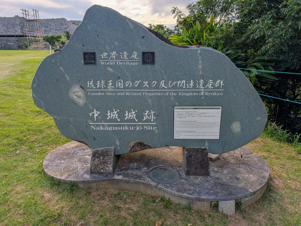 A stone sign on grass with information in Japanese and English about the World Heritage Gusuku Sites and Related Properties of the Kingdom of Ryukyu.