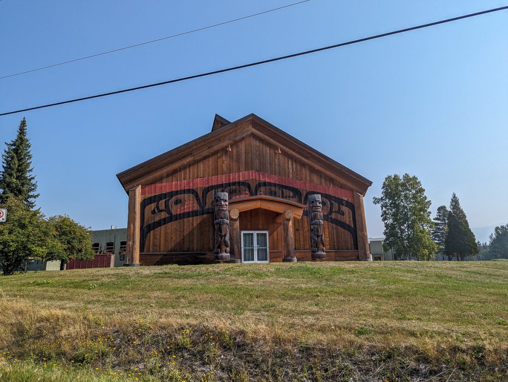 A wooden building with carved wooden totem poles flanking the doors, green grass in the foreground and a cloudless blue sky in the background.