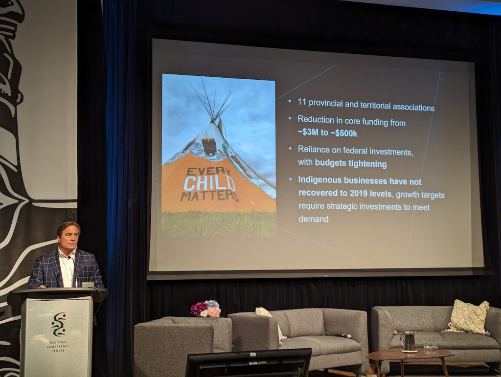 Keith Henry and Indigenous man and CEO, delivers a grim message at a conference. He stands at a podium with statistics on a projector. 