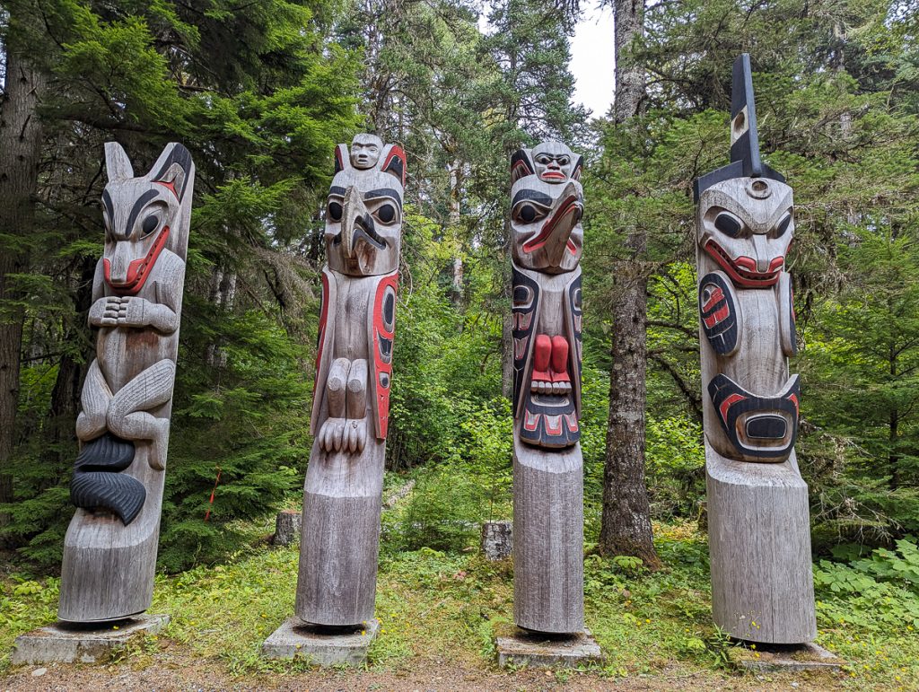 Four carved wooden totem poles with human/animal figures in a forest.